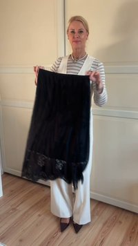 Μαύρο δαντελωτό φουλάρι που κρατιέται κατακόρυφα, με διακοσμητικό περίγραμμα με σχέδια. Το ύφασμα φαίνεται μαλακό με ρέουσα σχεδίαση. Λευκοί τοίχοι και ξύλινο πάτωμα βρίσκονται στο φόντο.