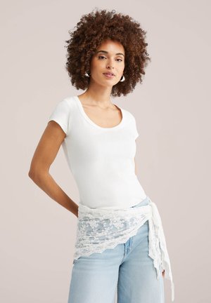 Kvinde med krøllet hår iført hvid t-shirt med korte ærmer, creol-øreringe, lyseblå jeans og et hvidt blonder tørklæde bundet om livet.