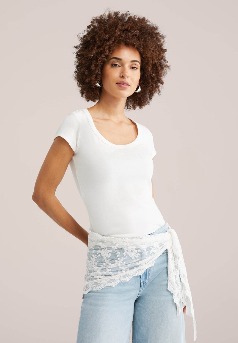 Femeie cu păr creț, purtând un top alb cu mâneci scurte, cercei cerc, blugi albastru deschis și un eșarfă albă dantelată legată în jurul taliei.