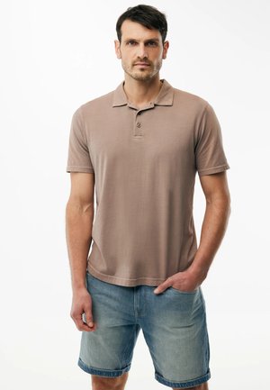 Mann mit kurzen dunklen Haaren, der ein hellbraunes kurzärmeliges Poloshirt und blaue, hochgekrempelte Jeansshorts trägt, steht mit einer Hand in der Tasche.