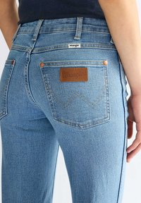 Άτομο που φορά ανοιχτό μπλε τζιν Wrangler με ορατές πίσω τσέπες, καφέ δερμάτινο λογότυπο και ραμμένο σχέδιο "W".