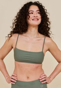 Donna con capelli ricci e scuri sorride, indossando un top e un bottom da bikini verde, in piedi con le mani sui fianchi contro uno sfondo beige.