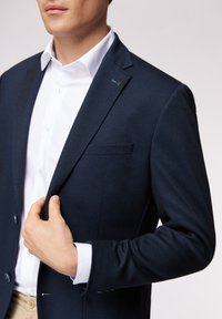 Granatowa marynarka o fakturze z klapami, pojedynczą kieszenią na piersi i niebieską przypinką. Nosi się ją na białej koszuli, prezentując coraz bardziej dopasowany fason.