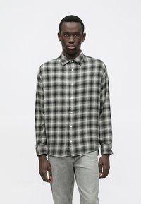 Νεαρός άνδρας που φοράει ένα μαύρο και άσπρο καρό πουκάμισο με μακριά μανίκια και κουμπιά, καθώς και ανοιχτό γκρι τζιν, στέκεται μπροστά σε λευκό φόντο.