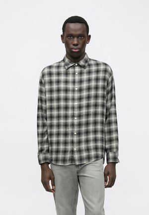 Junger Mann in einem schwarz-weißen karierten Langarmhemd mit Knöpfen und hellgrauen Jeans, der vor einem schlichten weißen Hintergrund steht.