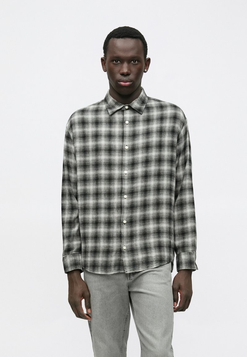 Νεαρός άνδρας που φοράει ένα μαύρο και άσπρο καρό πουκάμισο με μακριά μανίκια και κουμπιά, καθώς και ανοιχτό γκρι τζιν, στέκεται μπροστά σε λευκό φόντο.