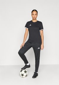 Μαύρο αθλητικό πουκάμισο με λευκές λεπτομέρειες και λογότυπο, σε συνδυασμό με μαύρο παντελόνι και ποδοσφαιρικά παπούτσια. Το μοντέλο στέκεται επάνω σε μια ποδοσφαιρική μπάλα.