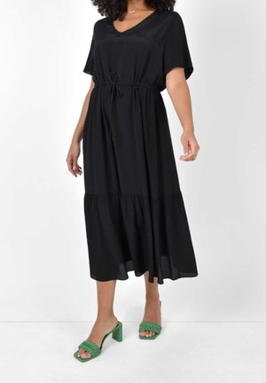 Abito midi nero con scollo a V, maniche corte, vita con coulisse e orlo a strati. Abbinato a sandali verde con tacco e cinturini intrecciati.