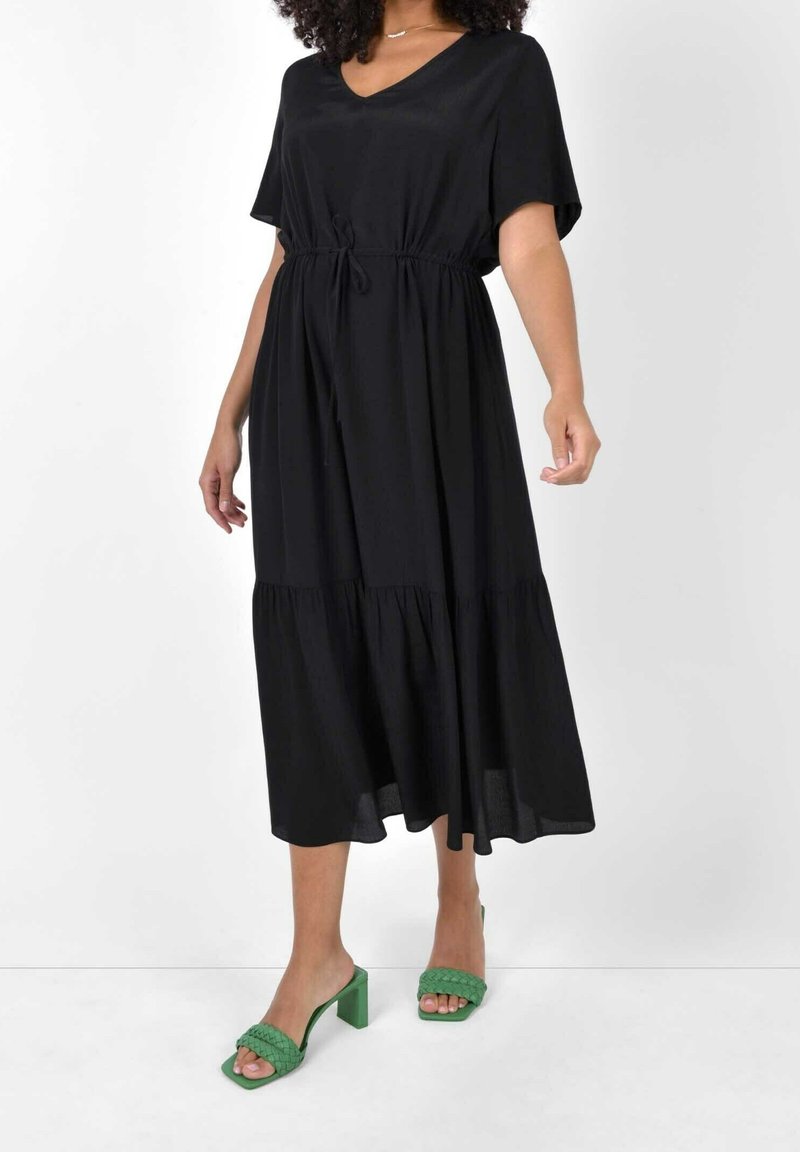 Svart midi-klänning med V-ringning, korta ärmar, dragsko i midjan och lagerkjol. Matchad med gröna klacksandaler med flätade remmar.