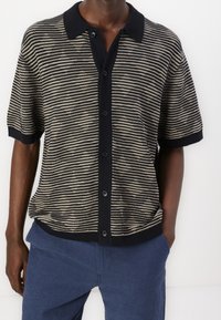 Mann in einem kurzärmligen, schwarz-beige gestreiften Hemd mit schwarzem Kragen und blauen Hosen, der vor einem weißen Hintergrund steht.