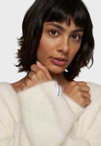Flauschiger weißer Pullover mit lockerer Passform und weiten Bündchen. Enthält ein Etikett am Ärmel. Der Hintergrund ist ein einheitliches Hellgrau.