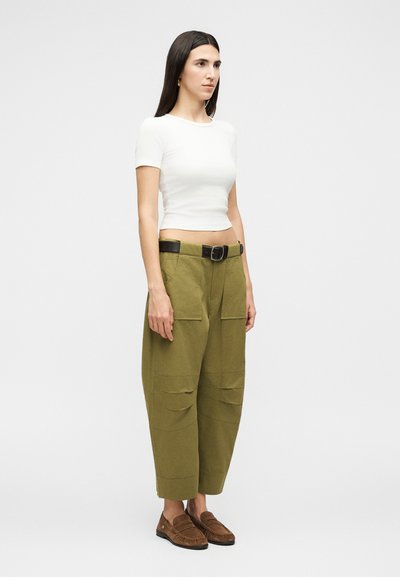 Γυναίκα με μακριά σκούρα μαλλιά, που φοράει άσπρο εφαρμοστό κοντομάνικο τοπ, ελαιοπράσινο παντελόνι με φαρδιά πόδια και τσέπες, μαύρη ζώνη και καφέ μοκασίνια, όρθια.