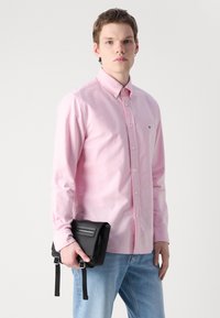 Rožā garām piedurknēm krekls ar pogu aizdari un apkakli, valkāts ar zilajiem džinsa bikšiem. Modelis tur melnu plecu somu ar siksnām un rāvējslēdzēju.