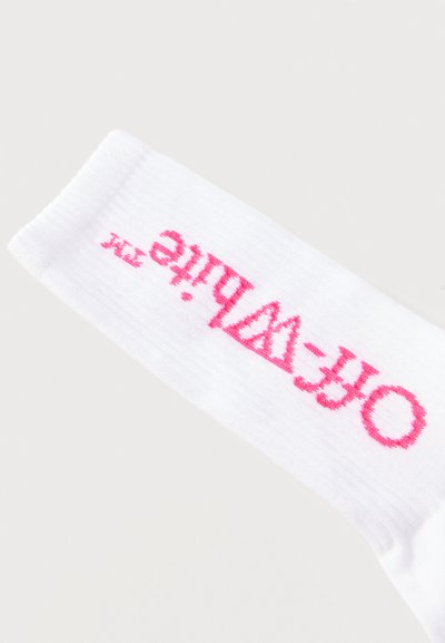 Chaussette blanche côtelée avec le texte rose « OFF-WHITE » tricoté horizontalement sur la partie du mollet sur un fond gris clair.