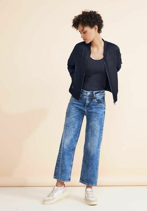 Dameoutfit med en sort bomberjakke, marineblå top, vide lyseblå jeans og hvide sneakers med pink detaljer. Enkel design.