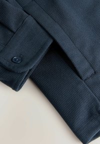 Un tessuto morbido di colore blu scuro presenta un design a coste con chiusura a bottone e dettagli sui polsini, evidenziando il suo aspetto curato.