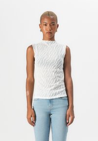 Hvit ermeløs topp med bølgete tekstur, høy hals og figurformet design, kombinert med lyseblå jeans. Glatt stoff og scallopede kanter.