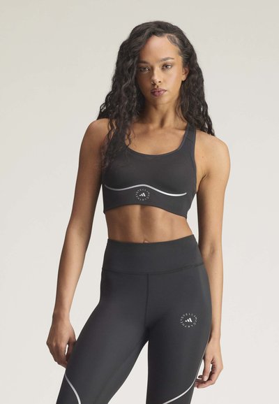 Ung kvinde iført sort sports-bh og leggings med reflekterende logo og detaljer, stående foran en ensartet baggrund.