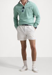 Pullover verde chiaro con colletto grigio, pantaloncini bianchi, sneakers bianche e calzini alla caviglia. Presenta un piccolo logo sul petto e occhiali da sole.