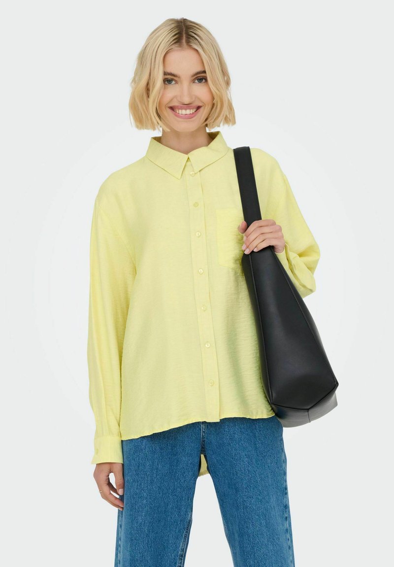 Hellgelbes Button-Up-Hemd mit einer Brusttasche, langen Ärmeln und einer lockeren Passform. Kombiniert mit blauen geraden Jeans und einer schwarzen Umhängetasche.