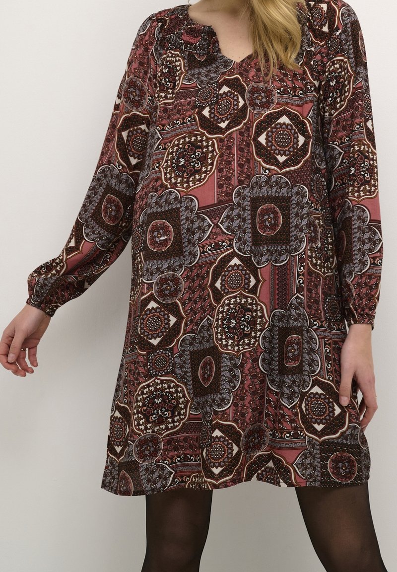 Knee-length šaty s dlouhým rukávem v vínové barvě, vyrobené z vzorované látky s květinovými a geometrickými vzory, se nařaseným výstřihem a uvolněným střihem.