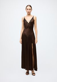 Brown satin maxi dress with deep V-neck and black lace trim, featuring a gathered skirt and side slit. Model wears black strappy heels.