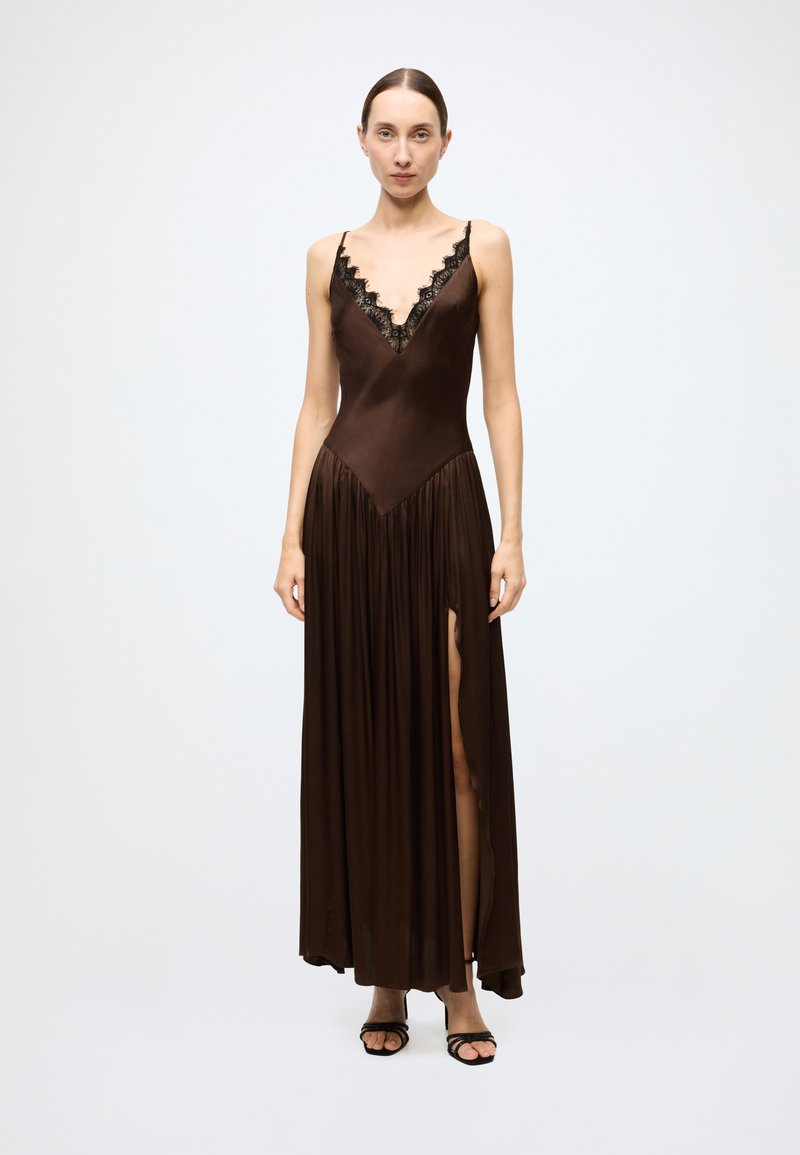 Brown satin maxi dress with deep V-neck and black lace trim, featuring a gathered skirt and side slit. Model wears black strappy heels.