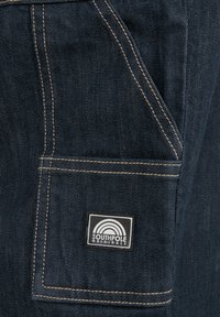 Taschina in denim scuro con cuciture in contrasto color tan. Presenta una patch rettangolare nera con un design a arco iris bianco e il testo "SOUTHPOLARIGINALS."