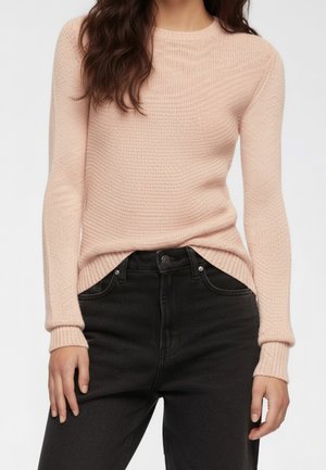 Maglione rosa chiaro lavorato a maglia con scollatura rotonda, polsini a costine e un motivo testurizzato, indossato sopra jeans neri a vita alta.