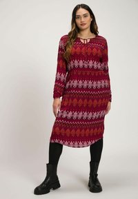 Langes rotes Kleid mit geometrischen Mustern in Weiß und Orange. Es hat lange Ärmel, einen runden Ausschnitt und wird mit schwarzen Stiefeletten kombiniert.