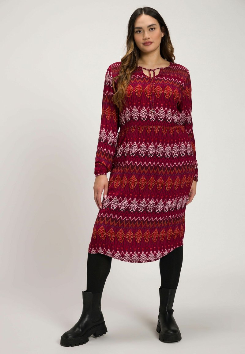 Langes rotes Kleid mit geometrischen Mustern in Weiß und Orange. Es hat lange Ärmel, einen runden Ausschnitt und wird mit schwarzen Stiefeletten kombiniert.