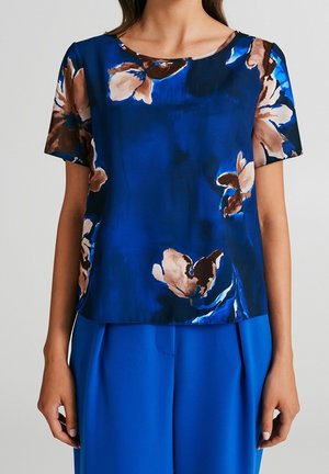 Kvinde iført en kortærmet, blå bluse med store brune og beige blomsterprint, kombineret med ensfarvede blå bukser.