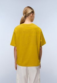 Femme avec une queue de cheval blonde portant un haut �à manches courtes jaune moutarde et un pantalon cargo blanc ample, se tenant devant un fond clair.