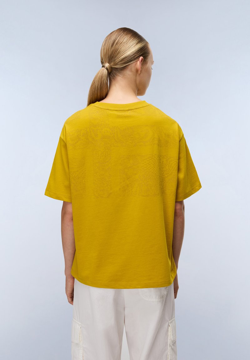 Femme avec une queue de cheval blonde portant un haut à manches courtes jaune moutarde et un pantalon cargo blanc ample, se tenant devant un fond clair.