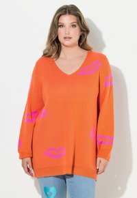Oranger Strickpullover mit V-Ausschnitt, verziert mit pinken Lippenabdruckmustern. Lange Ärmel und lockere Passform, kombiniert mit Jeans.