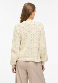 Femme aux longs cheveux bruns portant un pull en maille crème avec un motif texturé en forme de losange et un pantalon beige, vue de dos sur un fond blanc.