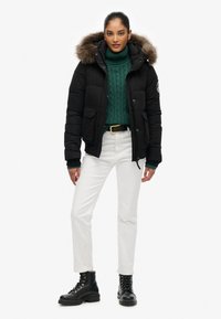 Sort dunjakke med pelsforet hætte, parret med en grøn kabelstrikket sweater, hvide bukser og sorte snørebåndsstøvler.