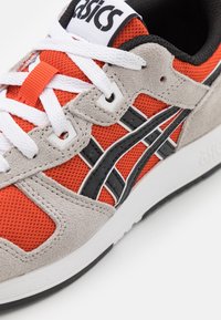 Scarpa sportiva rossa e grigia con tomaia in rete, sovrapposizioni in suede, strisce nere, lacci bianchi e suola strutturata. Logo sul tallone.