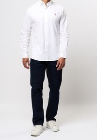 Biała koszula na guziki z długimi rękawami, zestawiona z ciemnoniebieskimi dżinsami i białymi trampkami. Na lewej piersi znajduje się małe logo.