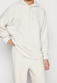 Persoon in een lichtbeige fleece hoodie en bijpassende sweatpants, met één hand op de heup en de andere ontspannen, tegen een effen achtergrond.