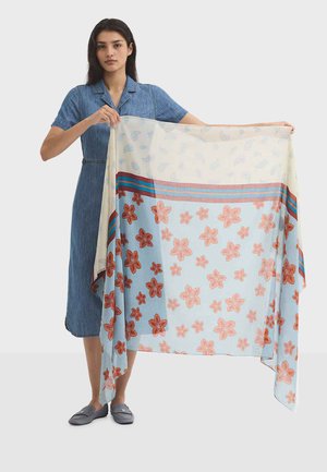 Femme en robe bleue tenant une grande écharpe florale avec des fleurs orange et des rayures bleues et rouges sur un fond clair.