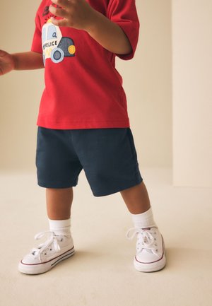 Peuter draagt een rood politie truckshirt, marineblauwe korte broek, witte sokken en witte sneakers, staand op een lichte vloer.