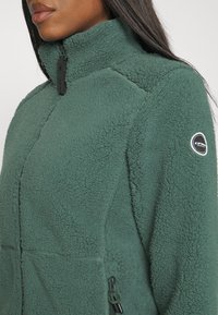 Giacca in pile verde con colletto alto, superficie testurizzata, tasche con zip e una patch con logo sulla manica. Presenta un design moderno.
