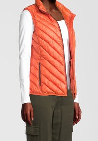 Pikowana pomarańczowa kamizelka z wysokim kołnierzem, z przednim zamkiem błyskawicznym i dwoma bocznymi kieszeniami. Noszona na białej koszuli z długim rękawem i zielonych spodniach.