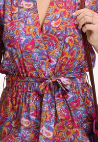 Színes ruha, amely élénk rózsaszín, narancssárga és kék virágminták keverékét kínálja; V-nyakú, meghúzott derék, kötővel, textúrált anyag.