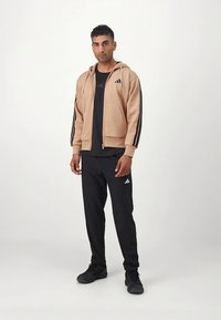 Sweatshirt com capuz castanho claro com fecho de correr e riscas pretas, combinado com uma t-shirt preta e calças de desporto pretas, todas com o logótipo da Adidas.