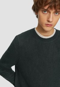 Dunkelgrüner gestrickter Pullover mit gerippter Textur und rundem Halsausschnitt, über einem weißen Hemd getragen, mit einer Nahaufnahme des Stoffes.