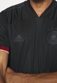 Maglia da calcio nera con tessuto testurizzato, design a collo a V e badge del paese in rilievo. Presenta dettagli rossi e dorati sulle maniche. Logo Adidas presente.