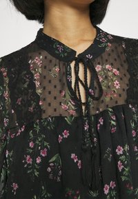 Blusa nera a fiori con un colletto in pizzo trasparente a pois, dettagli in pizzo scallopato e una chiusura con fiocco caratterizzata da nappine, che crea una texture stratificata.