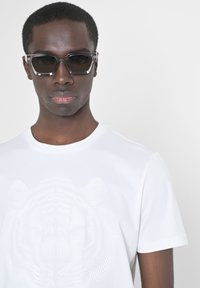 Zwarte rechthoekige zonnebril met dikke monturen en grijze glazen, gecombineerd met een wit T-shirt met een reliëfdesign van een tijger.